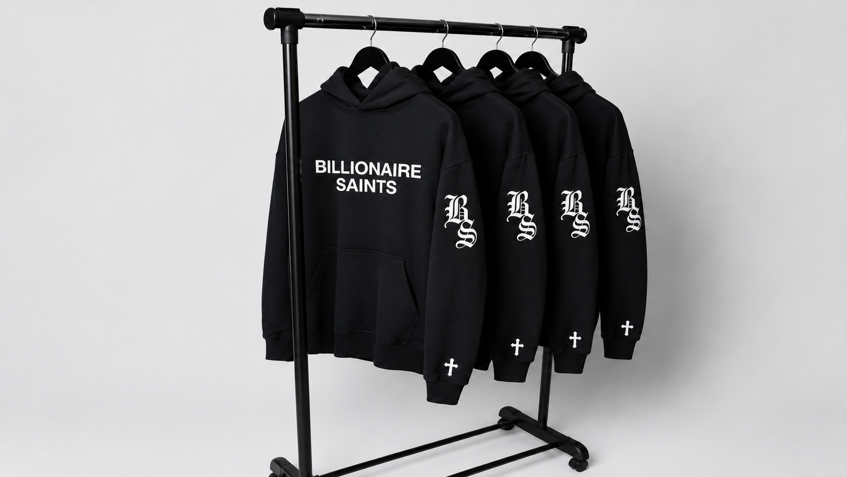A black clothing rack holding several black hoodies with white printed graphics. The front hoodie reads 'BILLIONAIRE SAINTS' across the chest, and the hoodies on the rack also show a large ornate 'BS' logo on the sleeve and a small cross near the cuff.